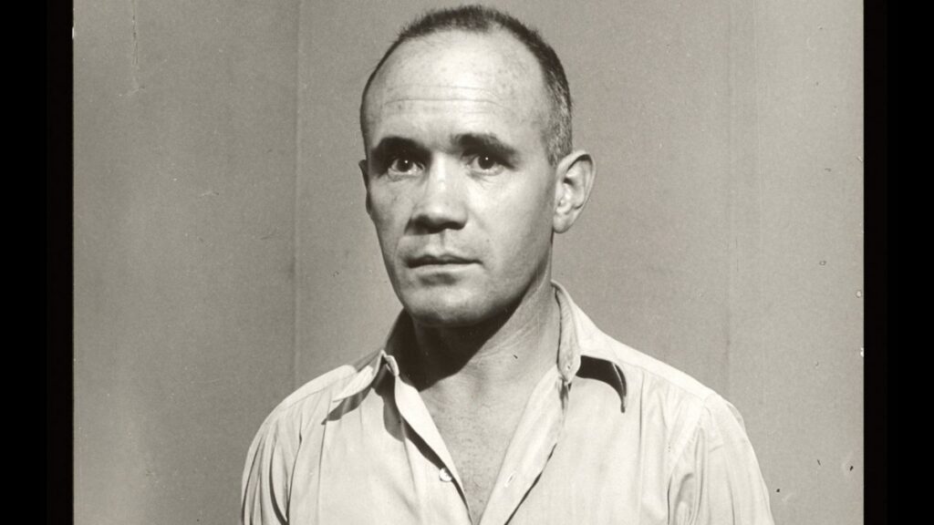 A black-and-white photo of a middle aged caucasian man (novelist, poet, playwright and filmmaker Jean Genet). He is looking into the camera. He is wearing a loose white shirt, has heavily lidded eyes, a wrinkled brow and receding hairline. His mouth is in a relaxed neutral expression.