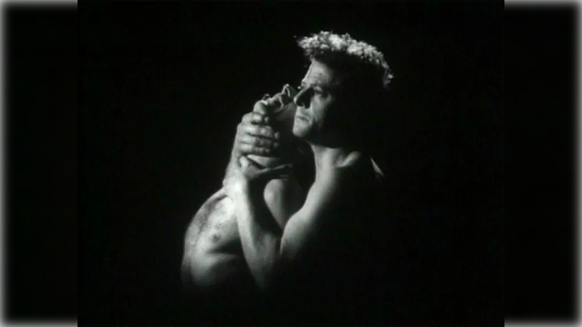 Against a completely black background. An older white man (the warden) caresses the throat of another male whose face is masked by the warden's hand. The photo is in black-and-white.