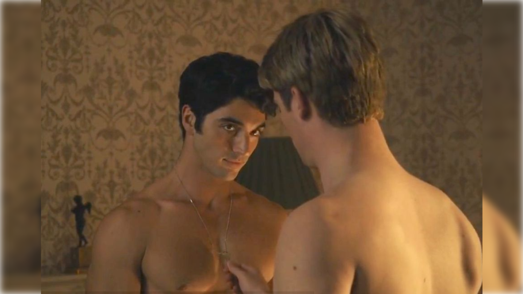 Prince Henry, a nude auburn haired Caucasian male caresses the chest of Alex, a nude dark haired Mexican-American male. Alex is wearing a house key around a chain and looking demurely into Prince Henry's eyes. Prince Henry has his back facing forward.