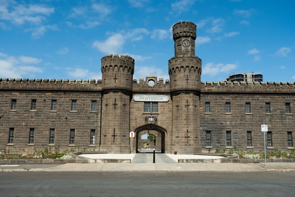 A stone prison entrance with two towers flanking the entry gate. In faded lettering is the words "Penal Establishment Pentridge". The picture is how the prison looks currently as both a museum and a residential village.