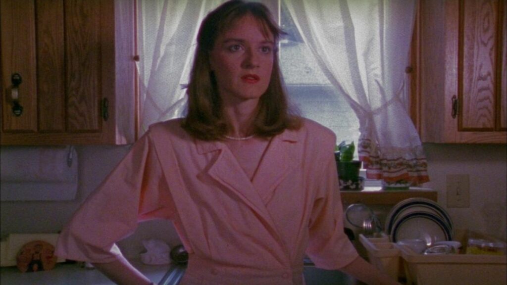 Felicia Beacon, played by Edith Meeks, stands in front of her kitchen sink in a pink dress with large 80s collar. Her hair is tidy and shoulder cropped of auburn tones. She looks exasperated and vacant.