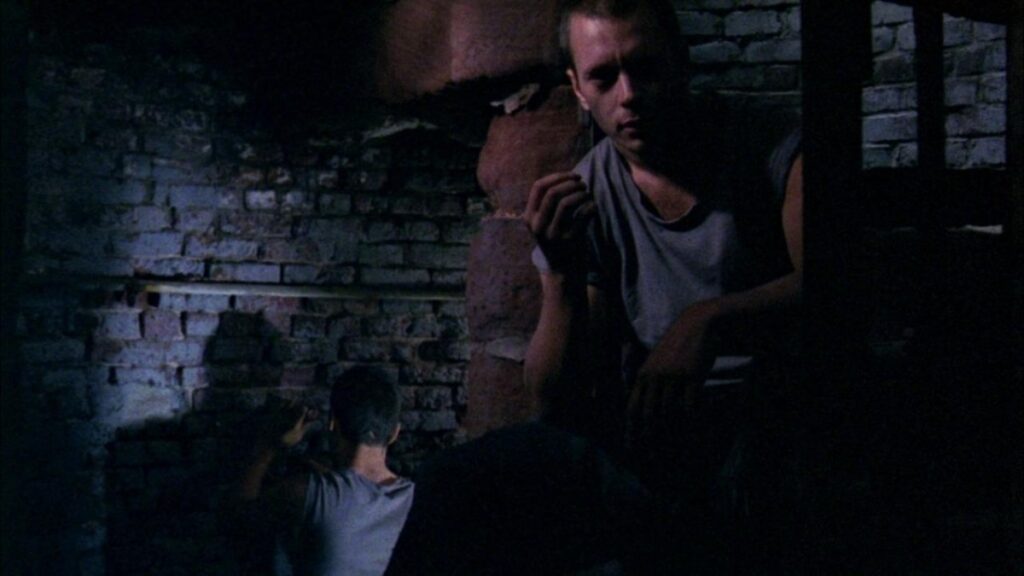 In shadowy darkness inmate Johnny Broom (Scott Renderer) looks towards the camera wearing a torn dark gray t-shirt against brick walls. He is sitting on the top bunk and holds a finished cigarette in his hand eith the back of a fellow inmate in the background of the shot.