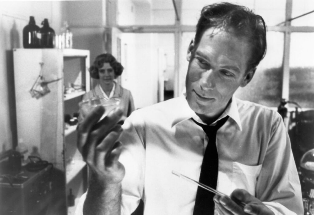 In black and white, Dr Thomas Graves played by Larry Maxwell investigates his "biomagnetic gas" after countless experiments. He is in the the foreground hold a small glass vesticle filled with dense white compound (something between liquid and gas) with a glass rod in his other hand. He is wearing a white shirt and black tie looses around his neck. In the background and out of focus, Dr Nancy Olsen (Susan Norman) approaches Dr Graves. She is an attractive woman with short blonde curly hair and a simple skirt suit outfit.
