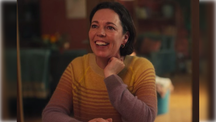 Olivia Colman, a caucasian brunette woman wearing a yellow jumper smiles to the camera.