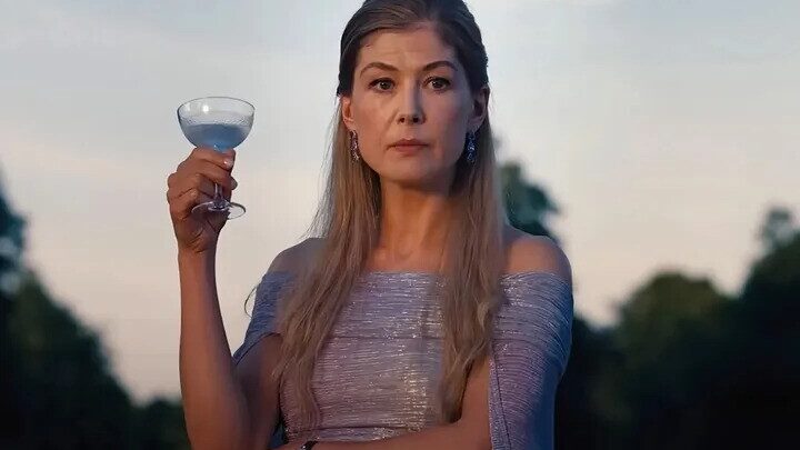Lady Elspeth Catton (Rosamund Pike) a tall willowy woman with straggly blonde hair is holding a champange saucer filled with a blue cocktail. She is wearing a pale lilac dress shod with silver.