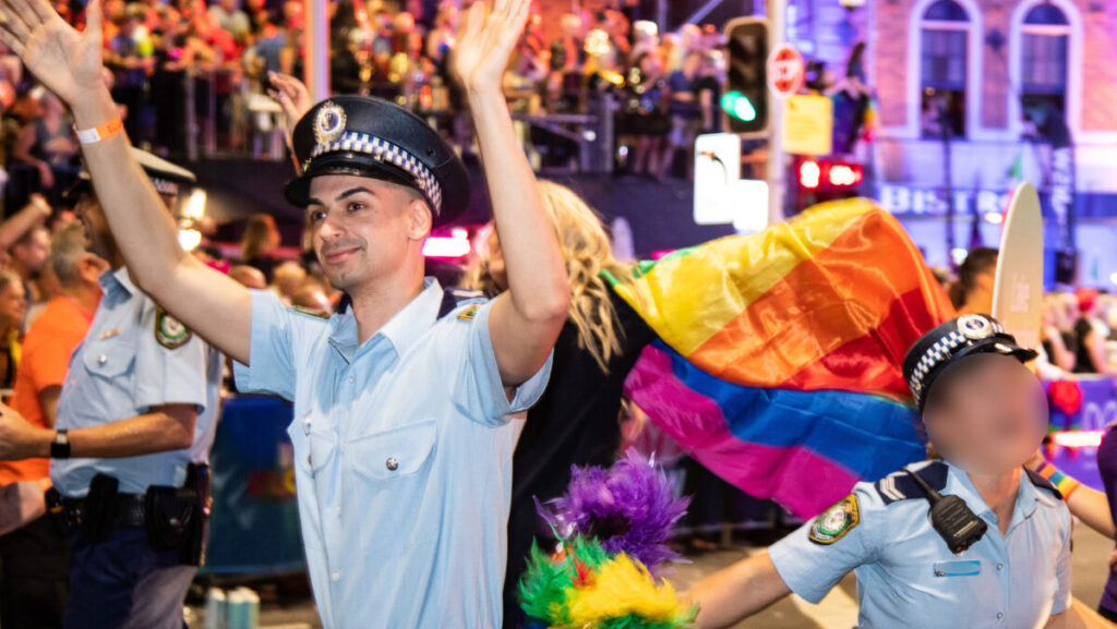 Senior Constable Beau Lamarr-Condon is seen in an Australian police uninform at the Sydney Mardi Gras parade in 2020. He is a thin male with olive skin. Behind him is a rainbow flag. Next to him another officer is seen celebrating however their face is blurred for privacy.