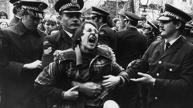 A person of indeterminate sex with glasses and a jacket is screaming while being grabbed from behind by a male police officer with a moustache in full NSW Policemans uniform. Another officer is helping him while in the background several other officers and activists are seen. The photograph is in black and white.