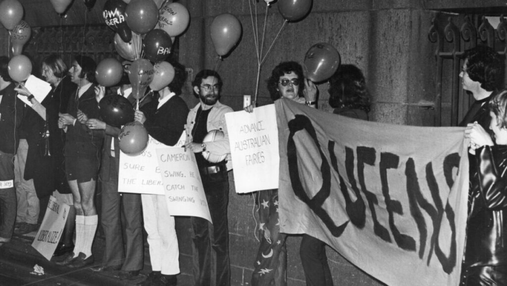 A line of homosexuals and lesbians holding bunches of balloons. One is holding a sign saying "Advance Australian Fairies" with two golding a banner saying "Queens". The photograph is in black and white.