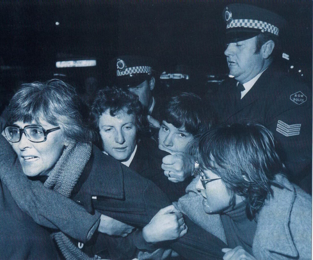 Four people are being grabbed aggresively by policemen. Gail Hewison in the foreground is wearing a woolen scarf, peacoat and sqaure rimmed glasses. A policeman is grabbing the shoulders of a person behind Gail of inderterminate sex. Gail's arm is being held by a fellow activist likely in an attempt to pretend her being pulled by the policeman, whose arm is grabbing Gail under her arms in the foreground. The photograph is in black and white.