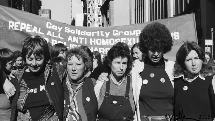 Five women are in the foreground wearing badges and t-shirts calling for Gay Liberation. In the background a large banner saying "Gay Solidarity Group: Repeal all Anti Homosexual Laws" is visible. There is further writing but it is unclear. The women in the foreground are described from left to right: A short dark haired woman with a black vest and flannel shirt over a black t-shirt saying "Gay M-"; an older woman with close cropped curly fair hair wearing a black vest, polo neck cardigan and a silk scarf; a middle aged woman with dark hair wearing overalls and a jumper; a taller woman with a dark large perm with a white cardigan over a black jumper and a long haired young man with a dark jumper and collared shirt. The photograph is in black and white.