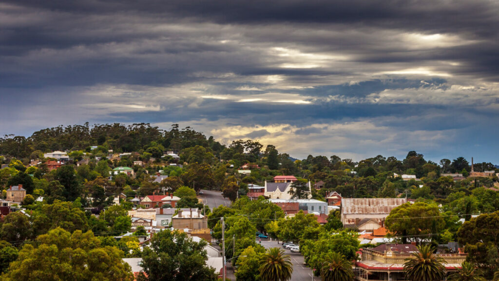 The town of Castlemaine stretches out with green hills and brick houses. Dark storm clouds sweep over the town but sunlight is still glinting through showcasing a hot wet summer.