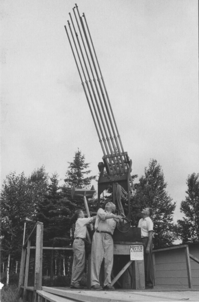 Wilhelm Reich, a middle aged man in a short sleeve shirt and trousers is turning the gear of a cloudbuster. The cloudbuster is a device with long thin tubes that point skywards. The photo is in black and white.