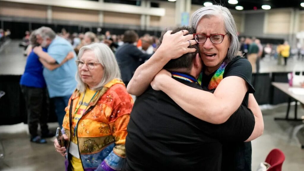 Two people are hugging and crying. The person facing their back to the camera is hugging a woman with a rainbow scarf. Behind them a shorter woman is standing wearing a colourful long-sleeved shirt.