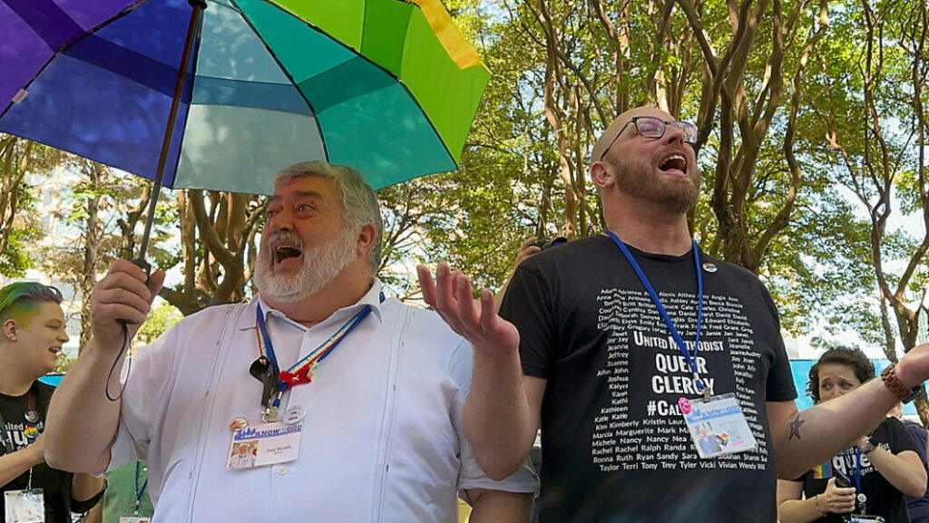 Two men are singing. The one on the left us bearded, portly and wears a white shirt. He is holding a rainbow umbrella in his right hand. To the right a bald bearded man with a black t-shirt has his hands open to the heavens.